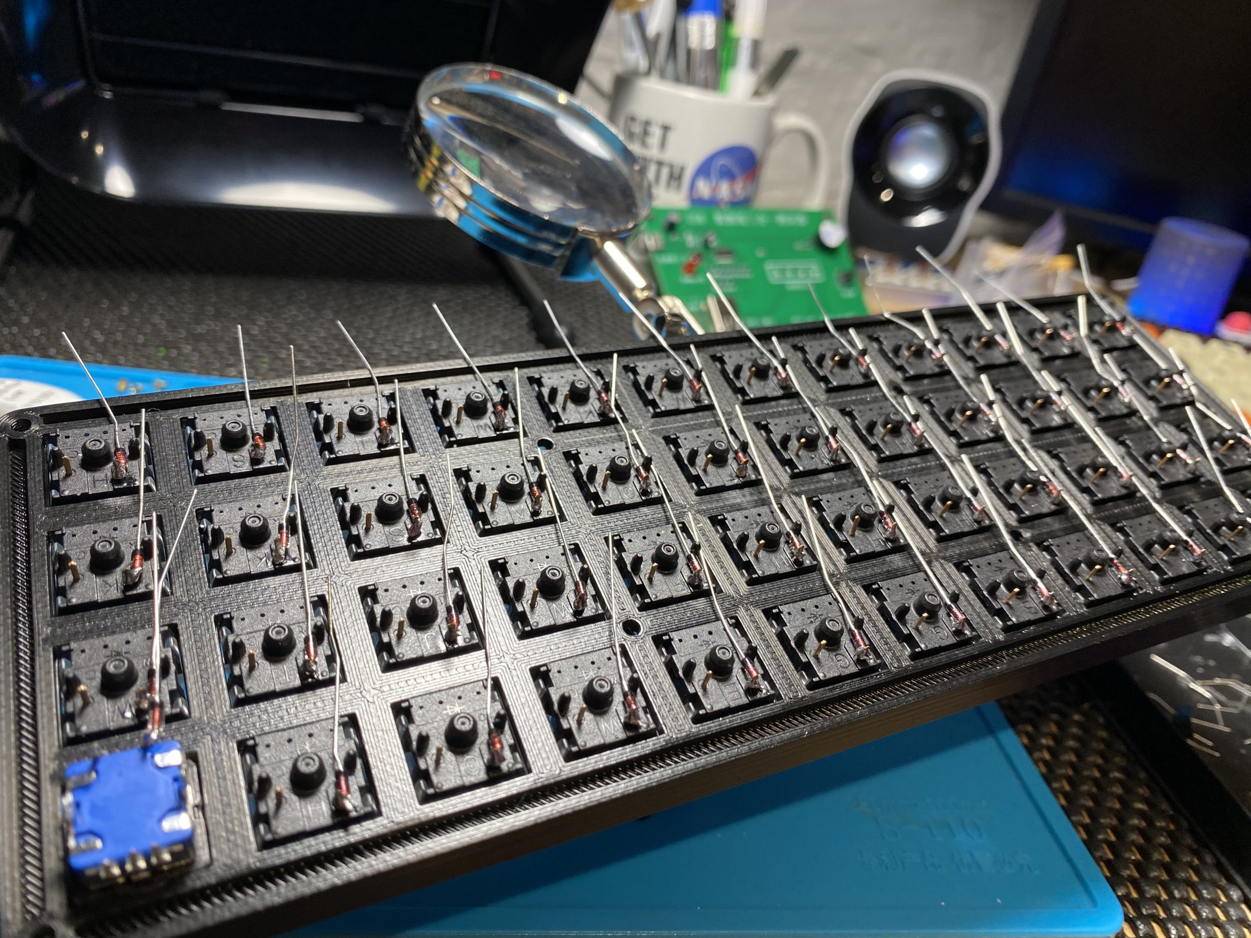 A zoomed out version of the previous picture, showing a 3d printed keyboard with all the switches inserted, viewed from the back, a diode has been soldered to each of the individual switches and a rotary encode is inserted in the top left most switch slot.