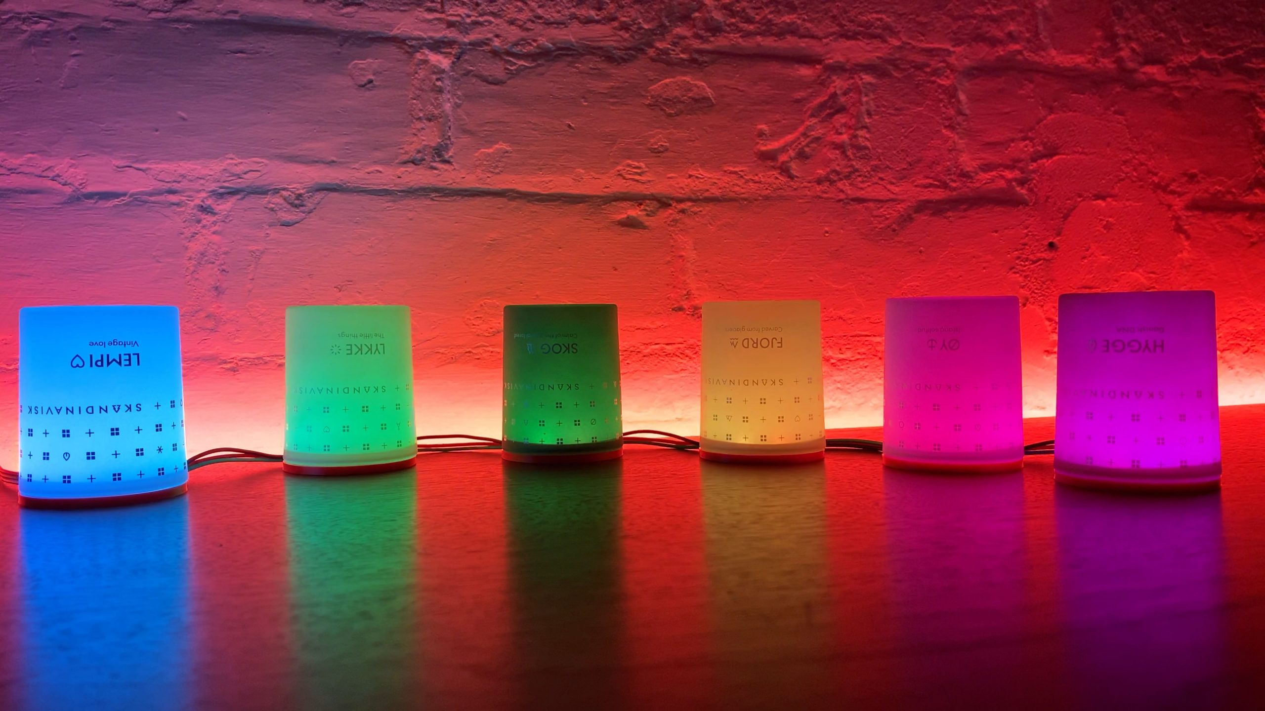 An image of six frosted glass candles with the wax removed, each about the size of a shot glass. The candles are upturned on plastic bases and lit up by leds in a rainbow colour, they are all sitting on a desk.