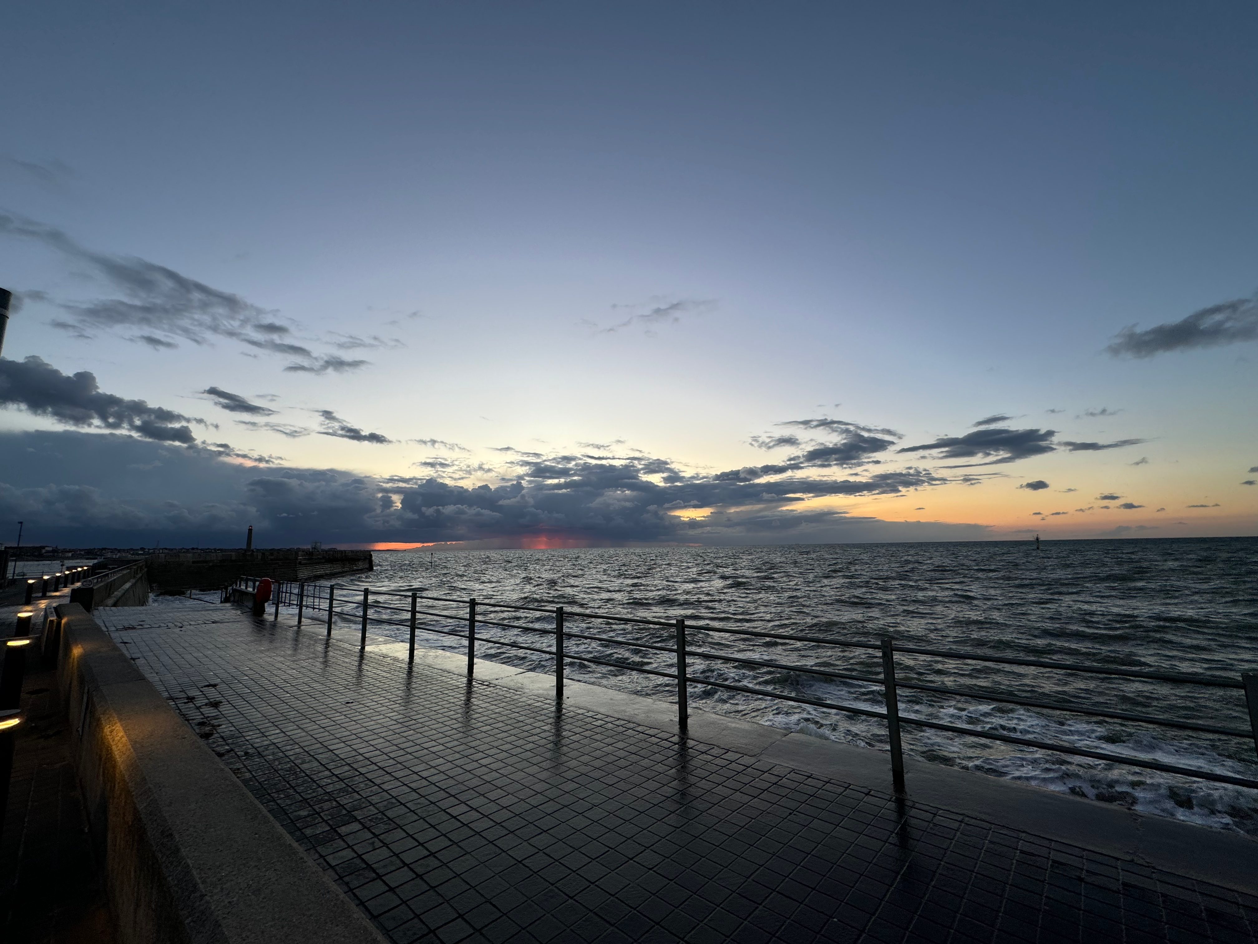 An image of a promenade by the sea at night, the picture is fisheye and it shows a turner sky (the artist) with moody clouds and choppy seas. There is a black looking metal barrier between the edge and the sea.